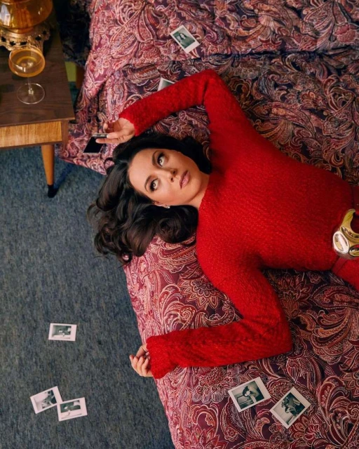 Aubrey Plaza lies on a bed in a red dress, surrounded by Polaroid photos; a side table holds a drink in a wine glass.