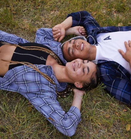 Two smiling people, a male and a female, lying in the grass in a field. They are both wearing casual clothing and appear relaxed and happy.