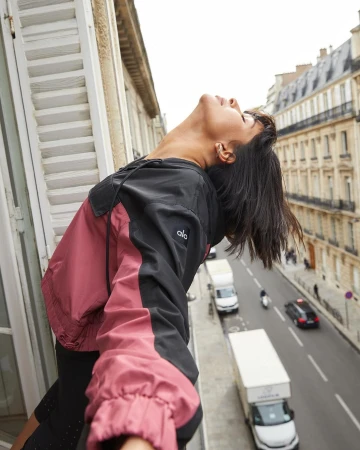 A woman in a pink and black jacket gazes upward while leaning out of a window with Paris buildings and street traffic visible in the background.