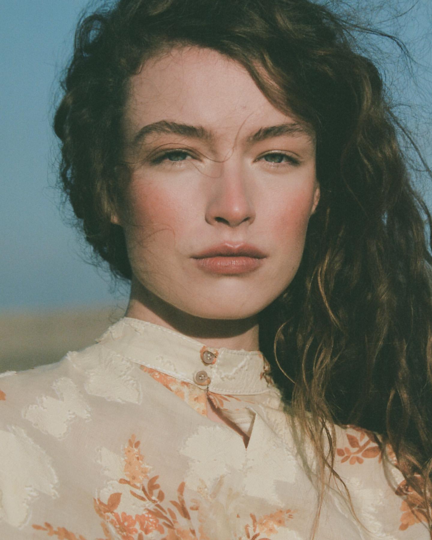 Close-up portrait of an adult female with long curly hair, wearing a floral-patterned high-neck top against a neutral outdoor backdrop.