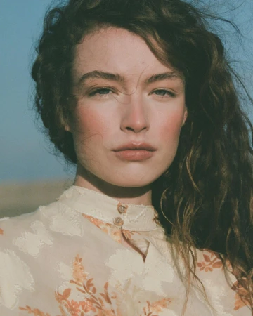 Close-up portrait of a woman with long curly brown hair wearing a patterned cream top against a clear sky background