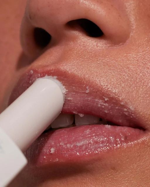 Close-up shot of an adult female applying lip scrub with a white tube applicator, lips slightly parted.