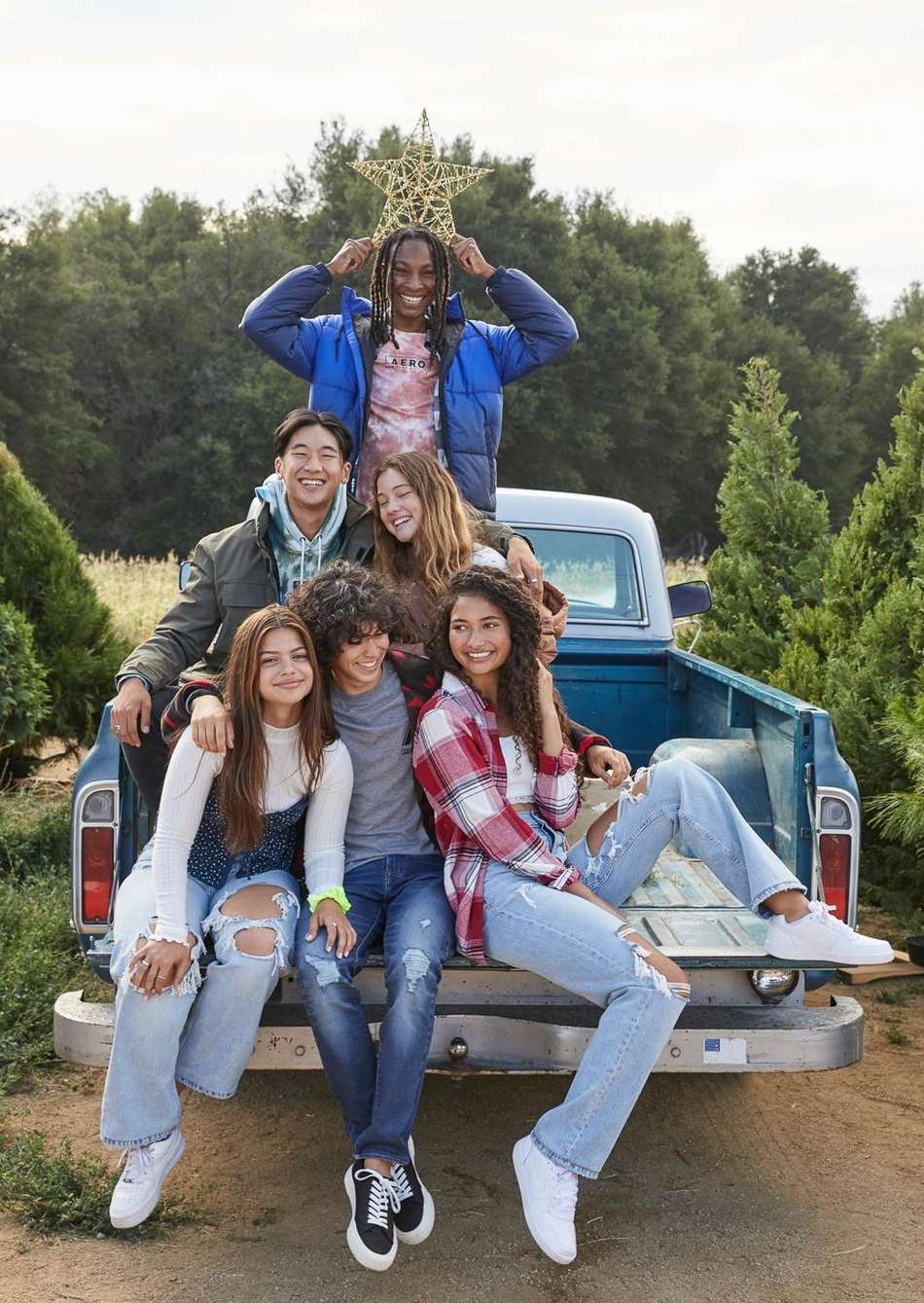 Group of six diverse teens sit on the tailgate of a blue pickup truck in a Christmas tree field on a sunny day, one holds a star.