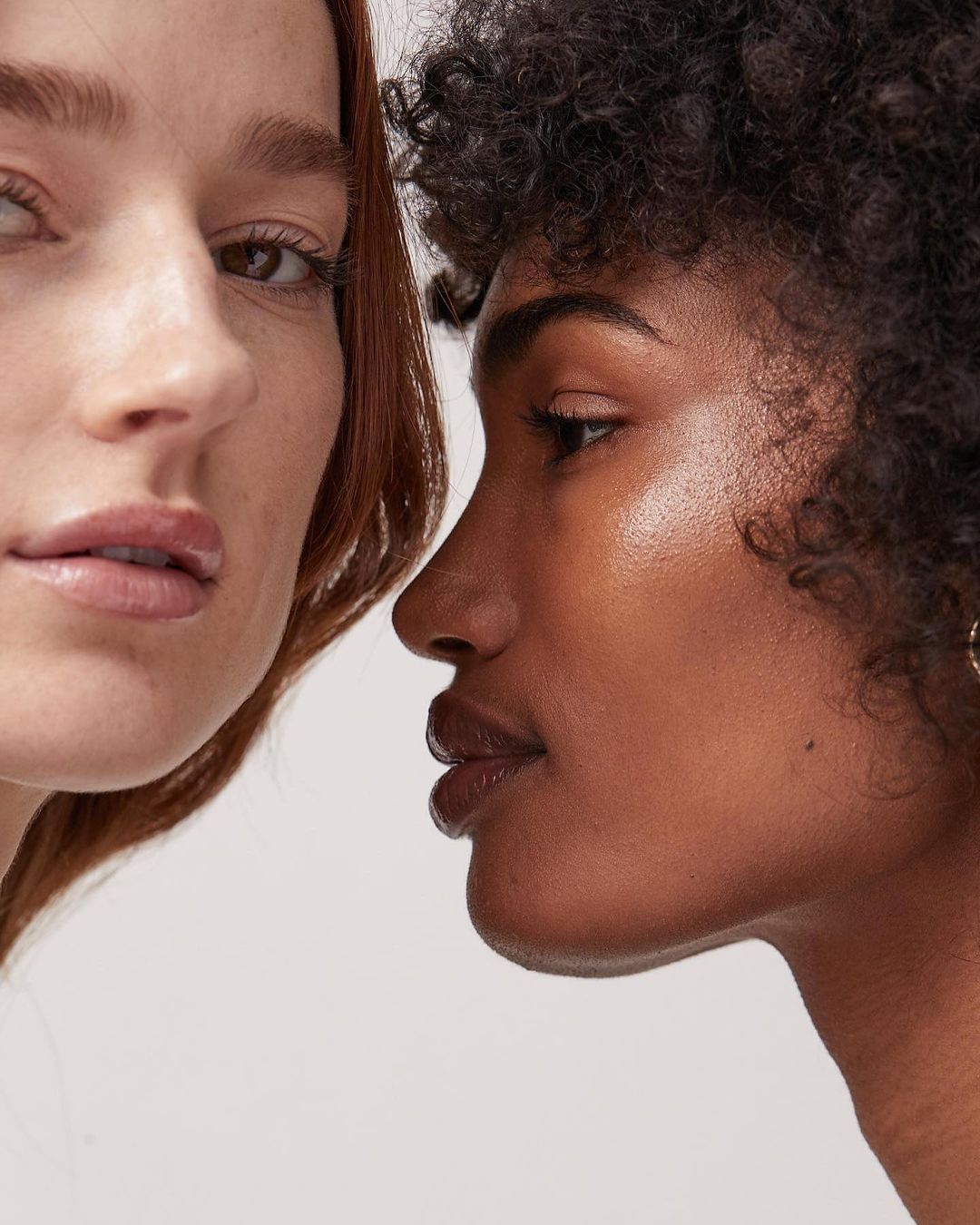 Close-up portrait of two women with contrasting skin tones in a beauty or skincare context, showcasing natural makeup and studio lighting.