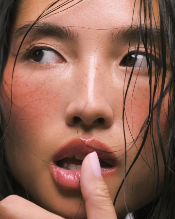 Close-up beauty portrait of a woman with wet dark hair, flushed cheeks, and freckles, touching her glossy lips with a pink-manicured finger