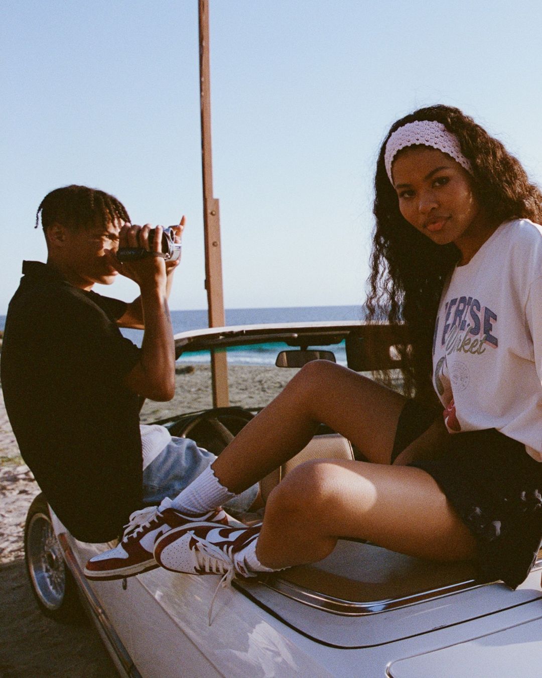 Two people in a white convertible car on a beach, one holding a vintage camera, under a clear sky with ocean in the background.
