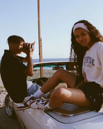 Two people in a white convertible car on a beach, one holding a vintage camera, under a clear sky with ocean in the background.