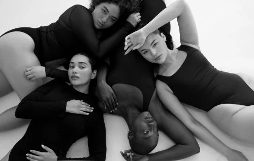 Four diverse women in black bodysuits pose on a white surface. A portrait format highlights the beauty and form of each individual.
