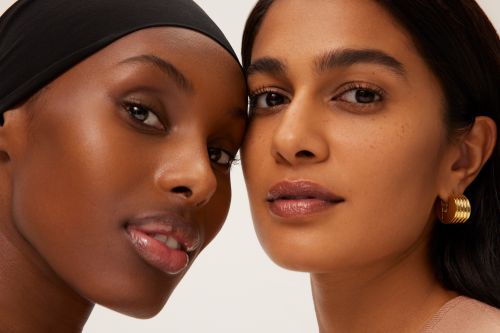 Close-up portrait of two diverse women with brown skin tones, radiant complexions, subtle makeup, and gold hoop earrings, set against a clean white backdrop.