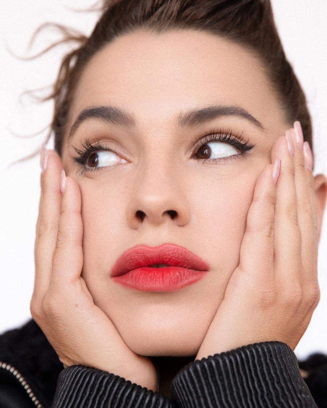 Portrait of a brunette woman with red lipstick, looking off-camera with hands framing her face against a clean, white background.