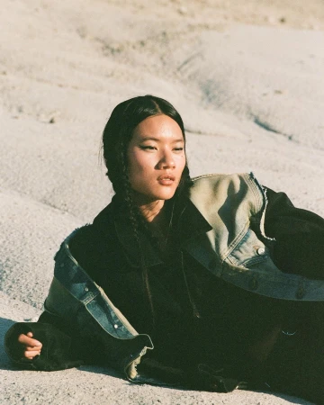 Model with braided dark hair wearing a denim jacket resting on sandy terrain under natural light