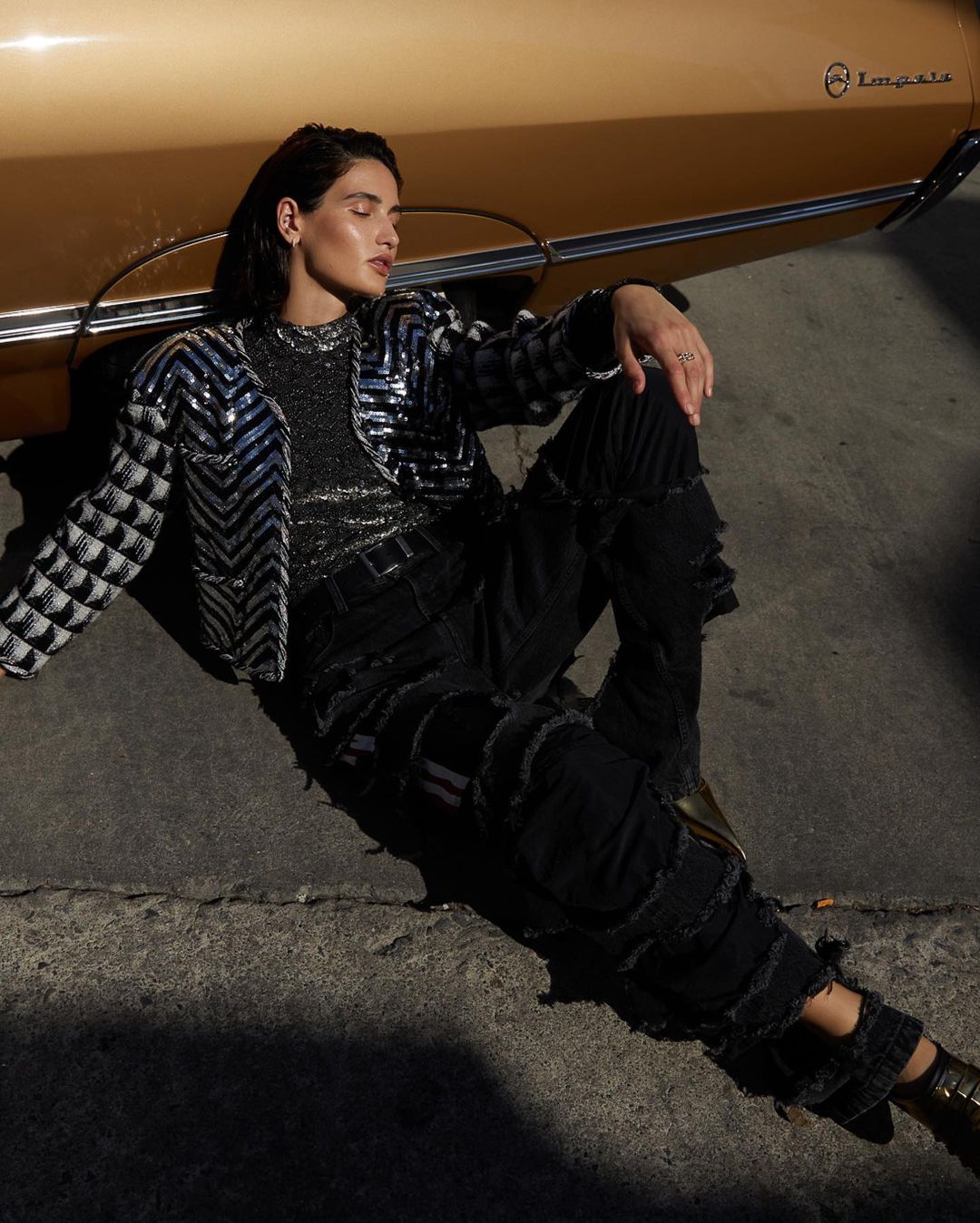Adult in stylish jacket and black jeans reclines on asphalt near a vintage gold car in outdoor fashion portrait; editorial style.