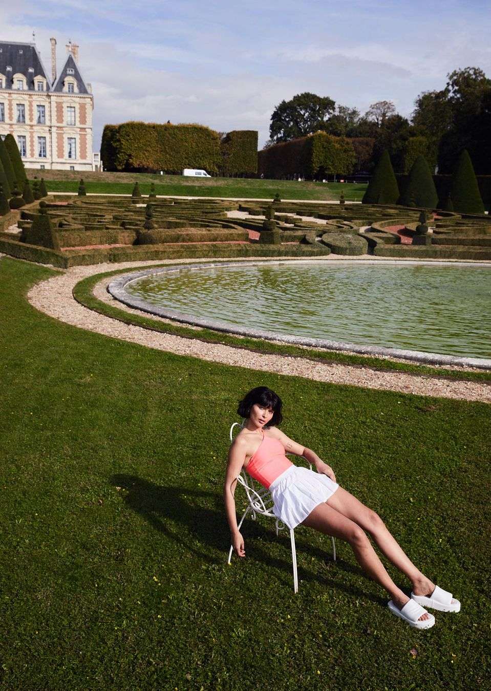 Young woman in an orange tank top and white skirt, sitting on a chair on green grass, with a chateau in the background on a sunny day.