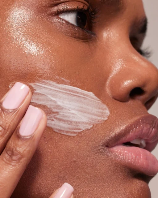 Close-up of a Black woman applying white cream to her face. Her pink fingernails are visible. Shot at eye-level in a studio.