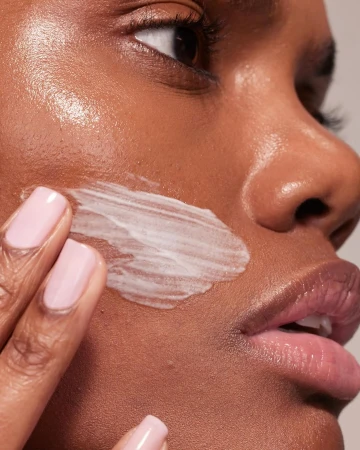 Close-up of a Black woman applying white cream to her face. Her pink fingernails are visible. Shot at eye-level in a studio.