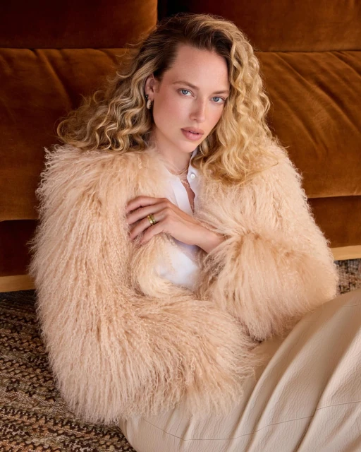 Portrait of a blonde woman wearing a shaggy jacket in a cream color indoors, sitting against a brown couch with a neutral colored rug.