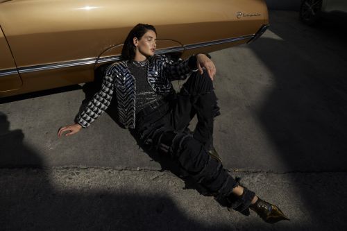 Adult woman in jacket and distressed black pants sits next to a vintage gold car on concrete in dappled sunlight in an outdoor fashion shot.