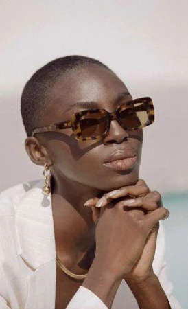 Portrait of a stylish short-haired black woman wearing patterned sunglasses and a white blazer against a blurred, neutral background.