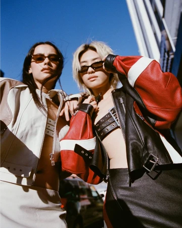 Two women pose in fashionable jackets against an urban background, showcasing modern outerwear and sunglasses in an outdoor setting.