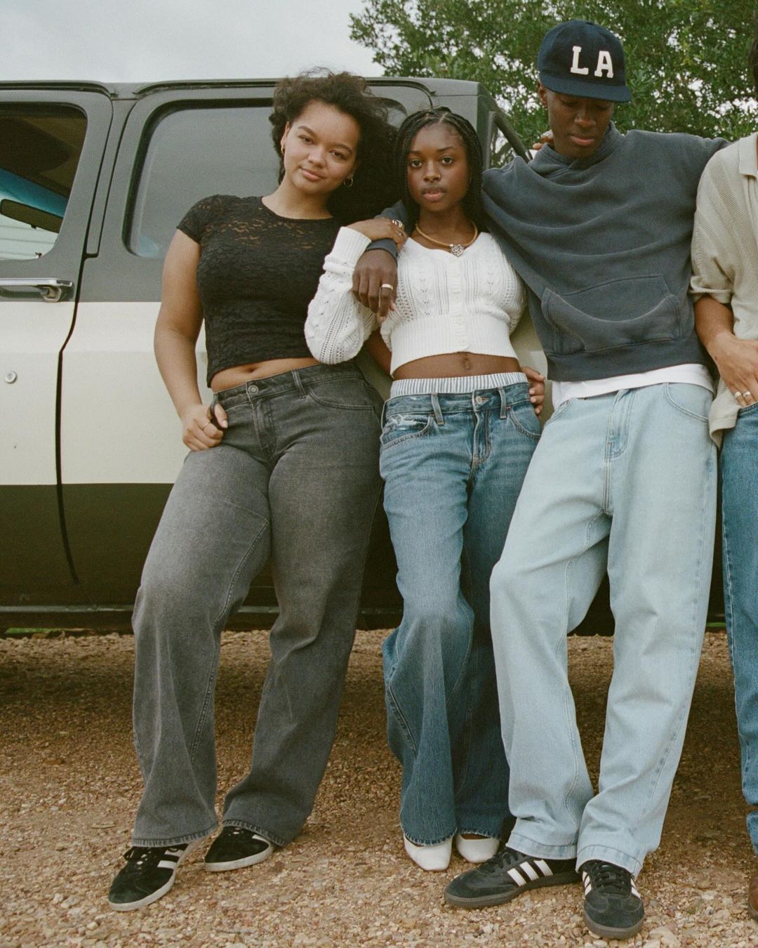 Group of young adults in casual fashion standing next to a vintage SUV, posing outdoors with natural lighting on a dirt road.