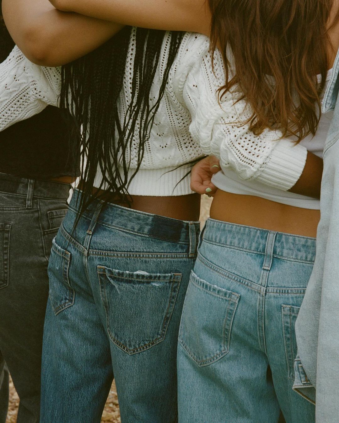 Three women embrace outdoors wearing cropped shirts and light wash jeans, showcasing modern fashion and natural skin tones in a lifestyle image.