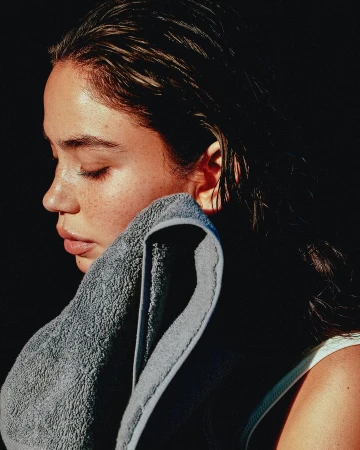 Profile portrait of a young woman with wet hair pressing a grey textured towel to her face against a dark background