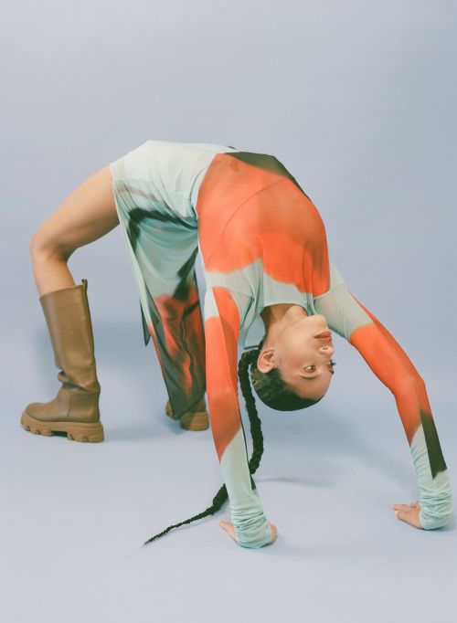 An adult woman in a patterned dress poses doing a backbend while wearing platform boots against a light blue backdrop in a studio shot.