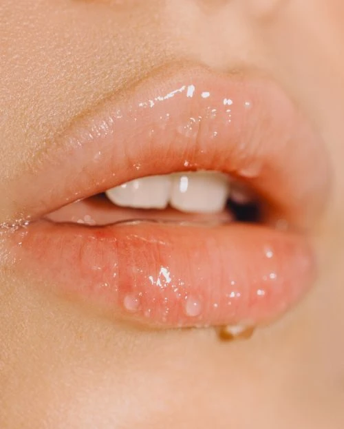 Extreme close-up of a woman's lips with glossy lip product, emphasizing texture and shimmer, set against a soft, out-of-focus background.