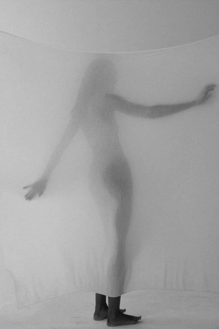 Monochromatic full shot of a figure behind a sheer white curtain, arms outstretched, showing the figure's outline in a minimalist studio setting.