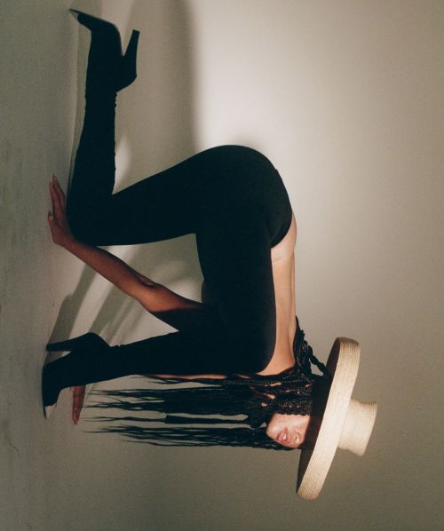 An adult female with long dark hair is upside down wearing black leggings, stiletto boots and a straw hat against a wall in studio lighting.