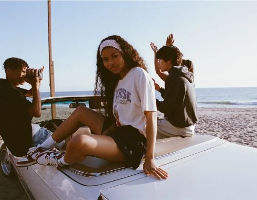 Three young people relax on a car at the beach on a sunny day; one takes a video while the others pose with ocean views.