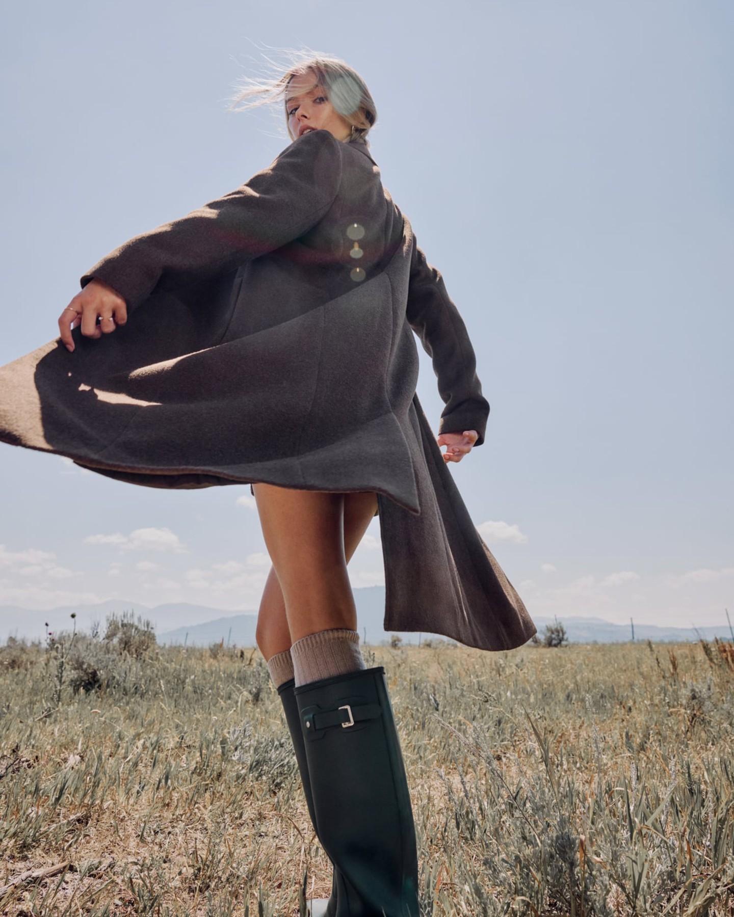 Young woman in coat and boots stands in sunny field with mountains in background against blue sky, looking over shoulder.