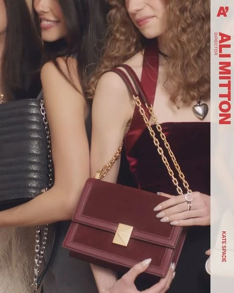Three women showcase Kate Spade handbags in a close-up shot. One wears a burgundy velvet top and gold jewelry, against a blurred background.