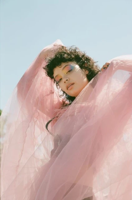 A young woman with dark curly hair stands outdoors under a blue sky, posing while draped with pink tulle fabric.