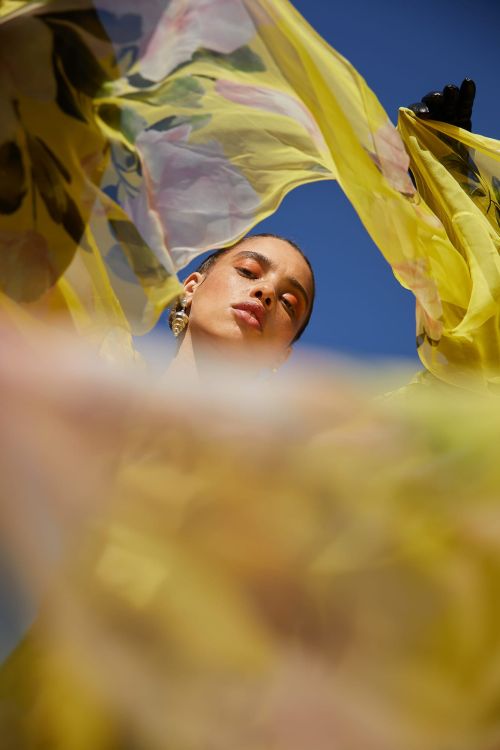 Woman posing with a yellow floral scarf against a blue sky. The scarf creates a frame around her face in an outdoor setting.