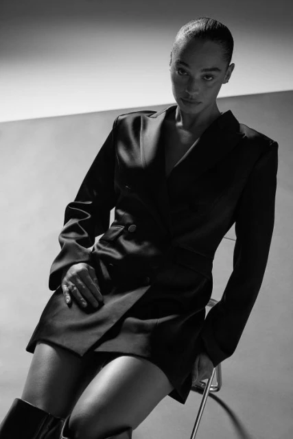 Elegant portrait of a Black woman in a black blazer dress, seated on a metal stool in a minimalist studio with soft, diffused lighting.