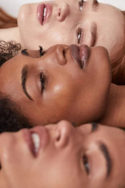 Close-up studio shot of three diverse young adult women lying in a row, focusing on their faces and glossy lips against a neutral backdrop.