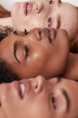 Close-up studio shot of three diverse young adult women lying in a row, focusing on their faces and glossy lips against a neutral backdrop.