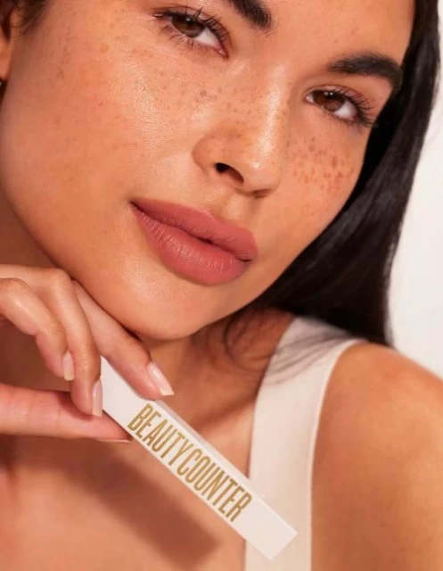 Close-up of a woman with neutral makeup, holding a Beautycounter product; tan skin, brown hair, and a focus on beauty and cosmetics.
