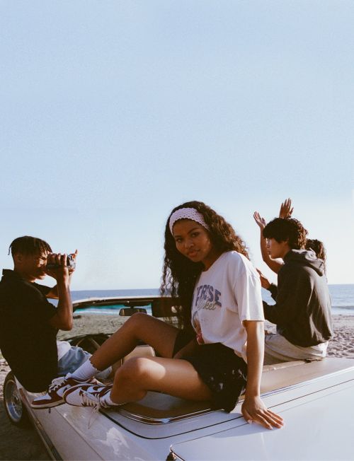 Young woman in a graphic t-shirt sitting on a convertible car at the beach with friends by Ali Mitton.