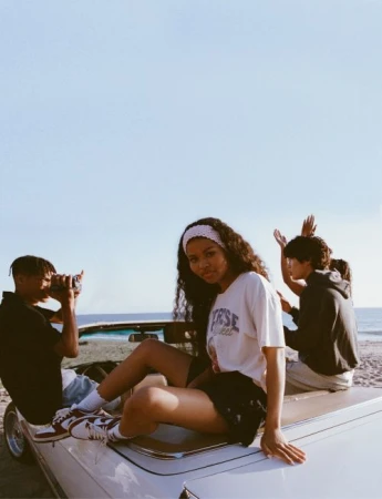 Young woman in a graphic t-shirt sitting on a convertible car at the beach with friends by Ali Mitton.