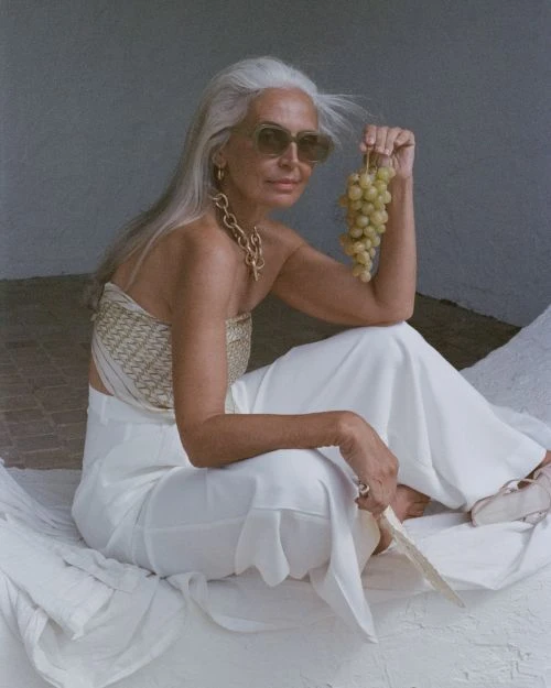 Mature woman with white hair poses with grapes. She is wearing a top, trousers, sunglasses, and jewelry. Shot in natural light.