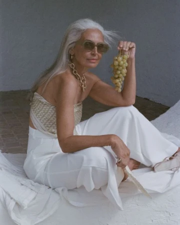 Mature woman with white hair poses with grapes. She is wearing a top, trousers, sunglasses, and jewelry. Shot in natural light.