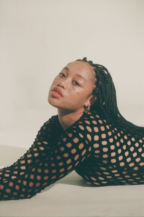 Portrait of a Black female model with freckles, wearing black fishnet top and gold hoop earrings, laying on a white surface.