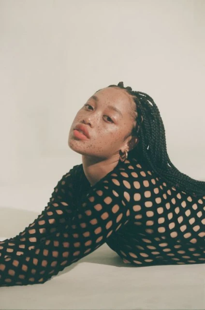 Portrait of a Black female model with freckles, wearing black fishnet top and gold hoop earrings, laying on a white surface.
