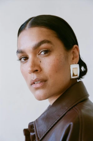 Close-up portrait of a woman with dark slicked-back hair wearing a brown leather jacket and chunky earrings against a white wall.