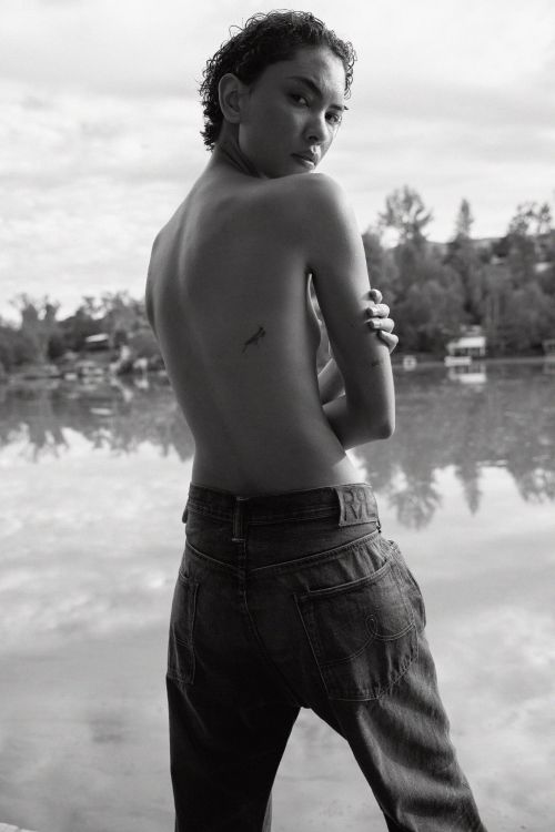 Adult posing outdoors in black and white, wearing jeans, with short hair, looking over her shoulder, with a body tattoo.