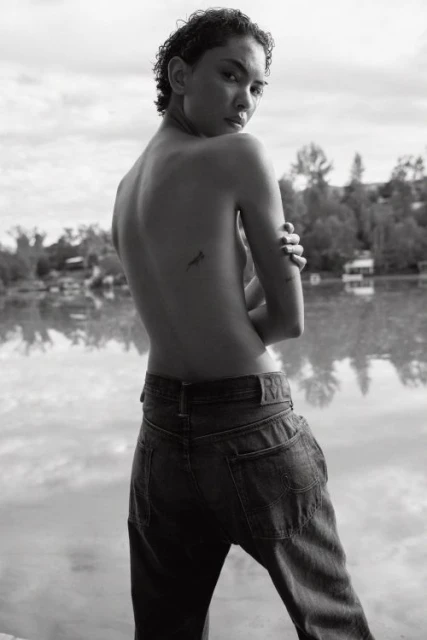 Adult posing outdoors in black and white, wearing jeans, with short hair, looking over her shoulder, with a body tattoo.