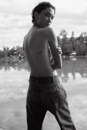Adult posing outdoors in black and white, wearing jeans, with short hair, looking over her shoulder, with a body tattoo.