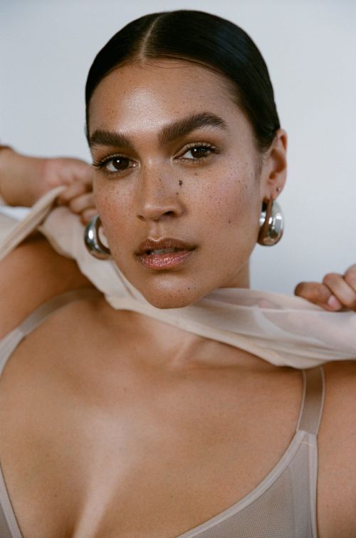 Close-up portrait of a woman with dark hair, fair skin, hoop earrings, and a beige top, posing against a neutral background in soft lighting.